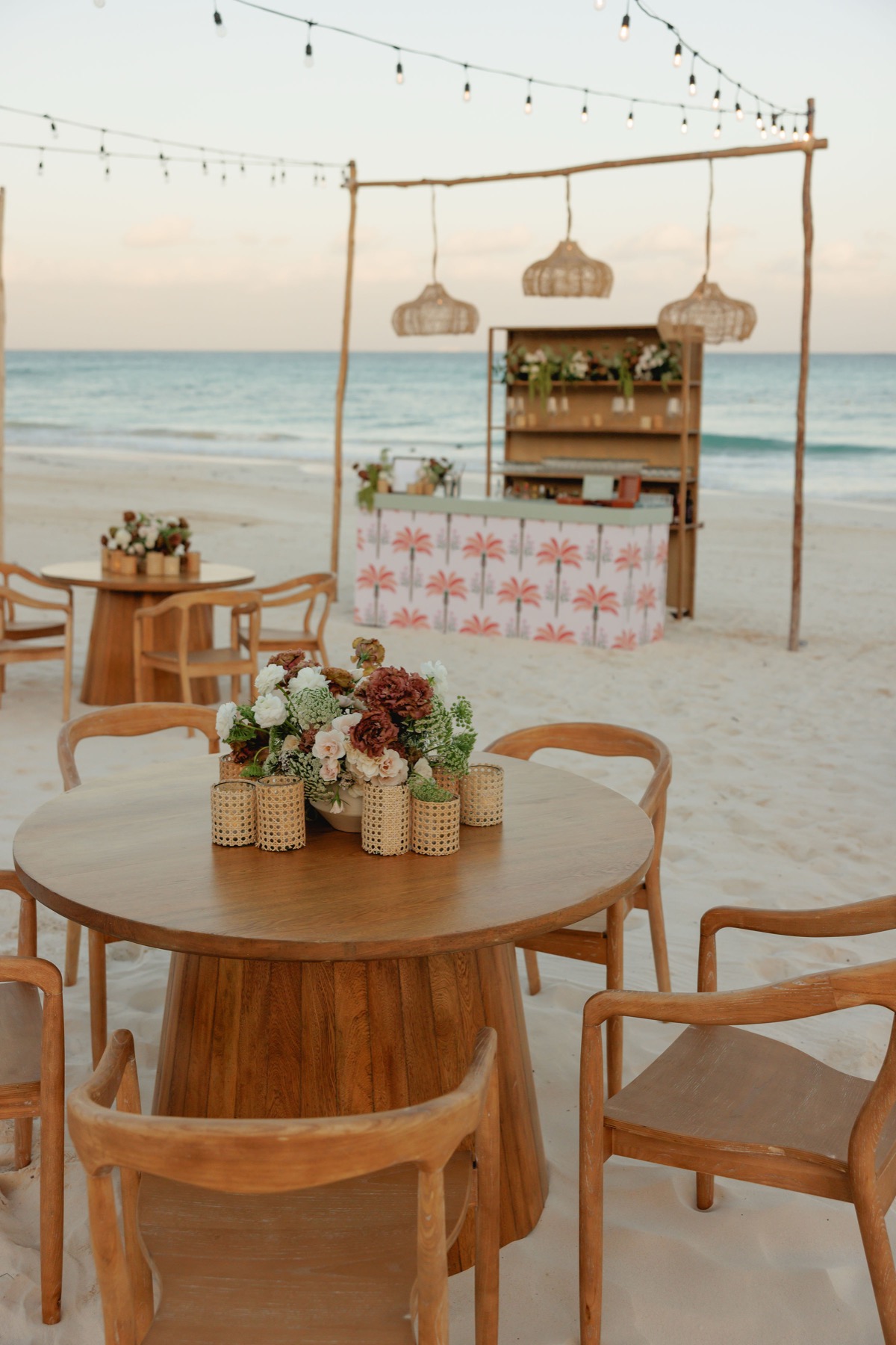 Welcome cocktail setup on the beach with a floral bar and round wooden tables at dusk