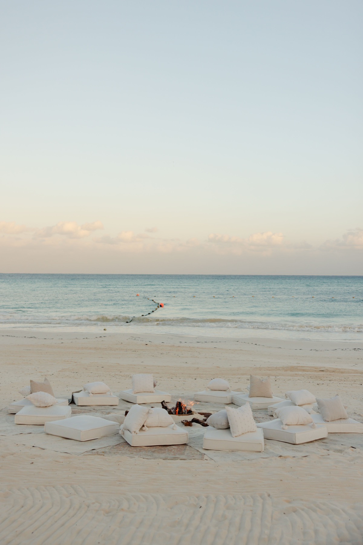 Beach lounge with white cushions around a fire pit, ocean view at sunset