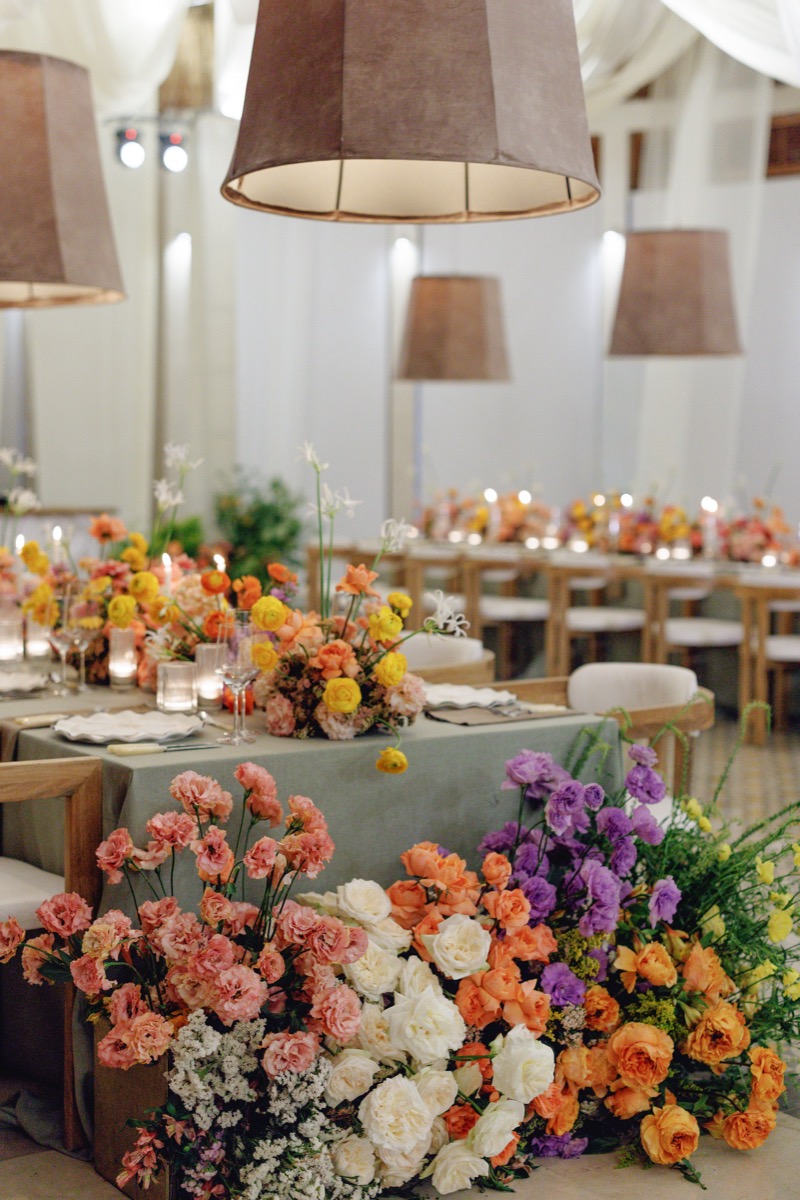 Reception tables with abundant orange, coral and violet florals under brown pendant lamps