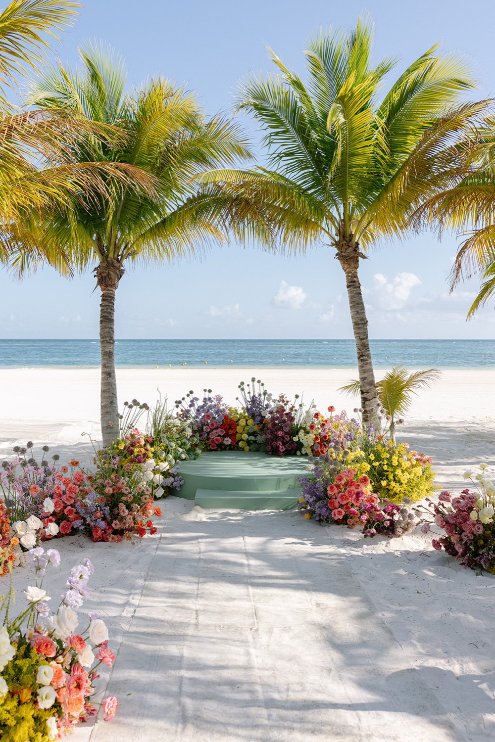 Beach ceremony altar between two tall palms, with a stage of low colorful florals facing the Caribbean