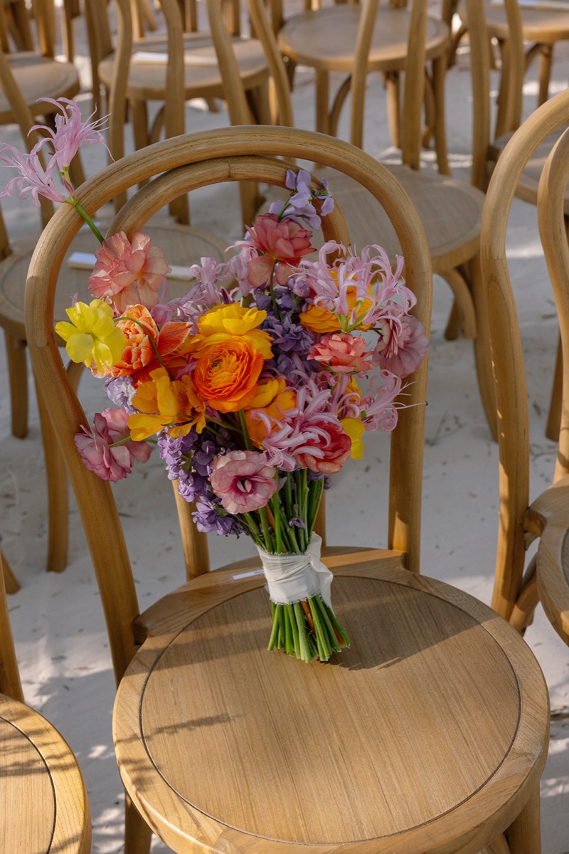 Hand-tied bridal bouquet of pink, orange, yellow and violet blooms resting on a cane chair