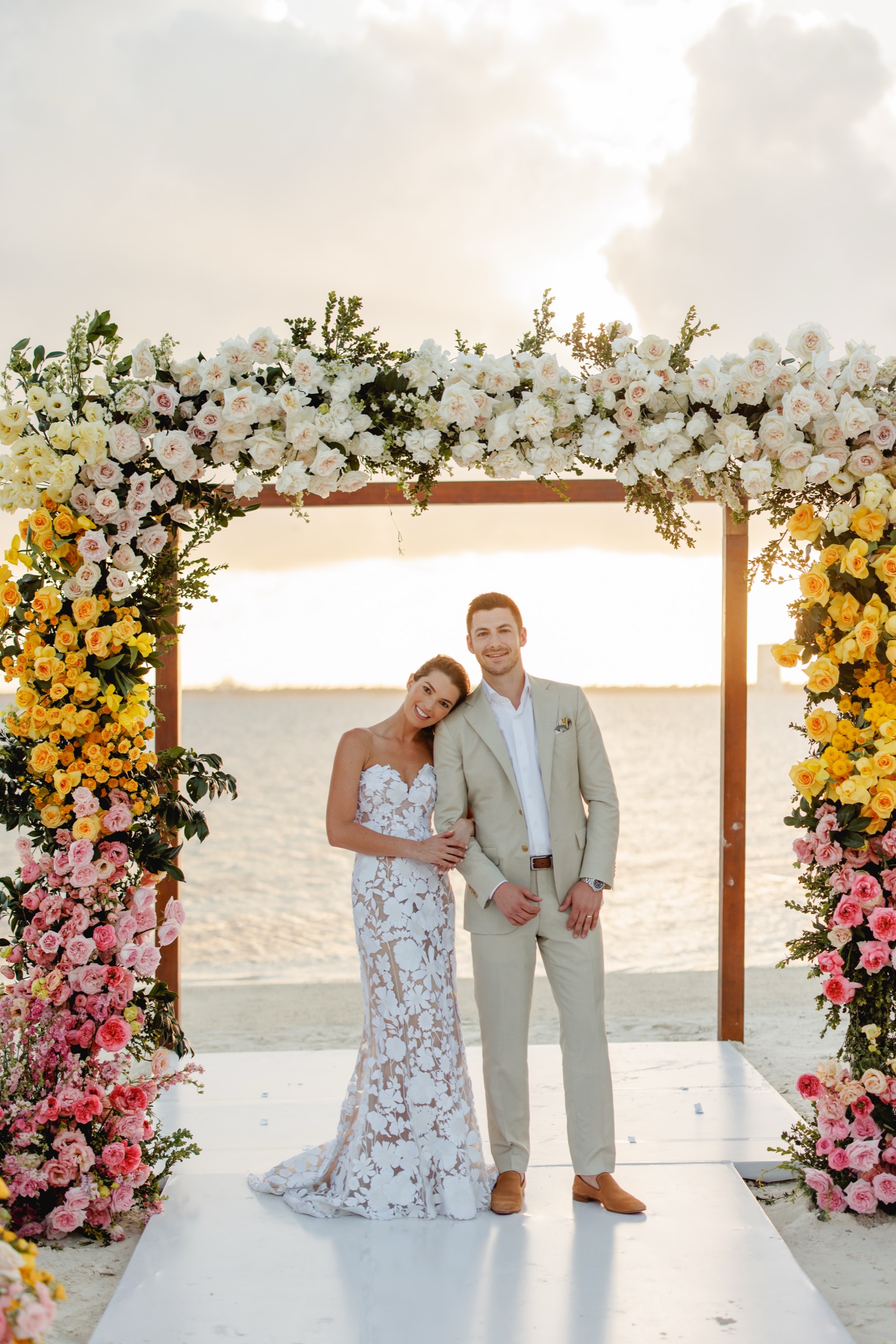Couple under a sunset floral arch designed by Canteiro